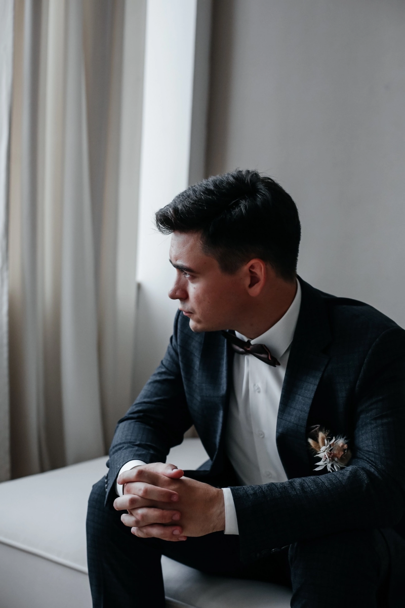 A man in a suit with a bow tie sits with his hands clasped together, looking out the window.