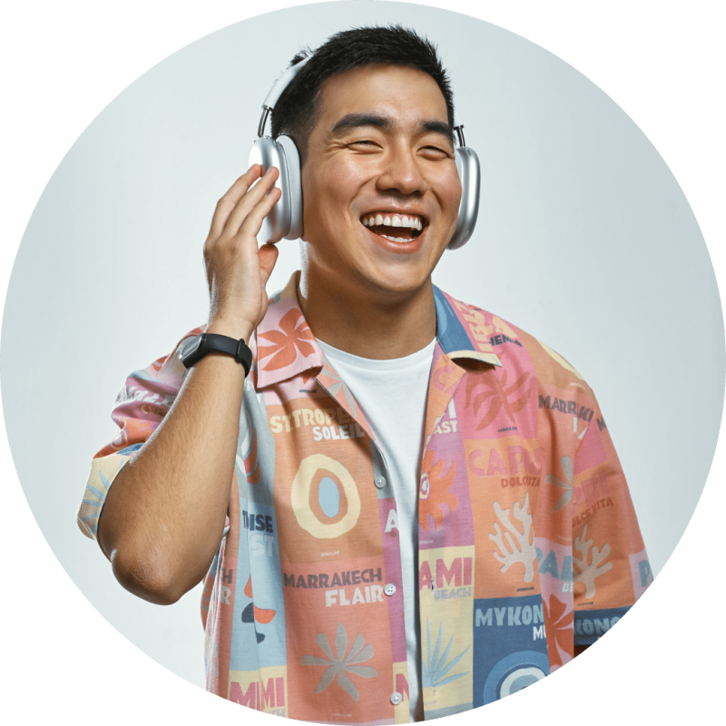 A smiling Asian man wearing silver headphones and a colorful shirt with various words such as 'Marrakech Flair' and 'Mykonos' is visible.