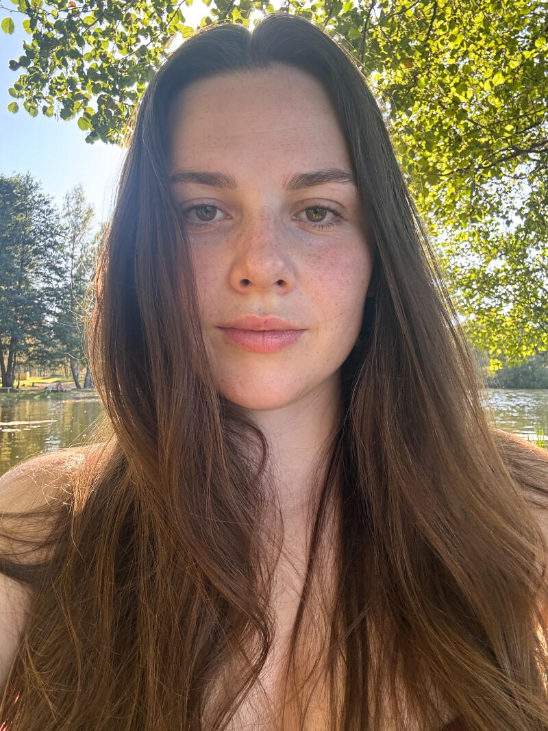 A close-up shot of a fair-skinned woman with long brown hair and freckles, standing outside near a lake and trees.