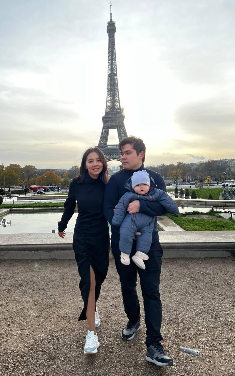 A family of three poses in front of the Eiffel Tower; the man holds a baby bundled in a blue snowsuit, while the woman wears a black dress and white sneakers.