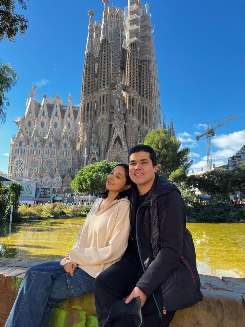 A couple poses in front of the Sagrada Familia in Barcelona on a sunny day. The woman wears a beige sweater and jeans, while the man wears a black jacket and pants.