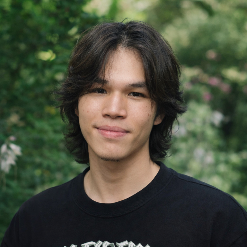 A portrait of a young man with dark hair and a black shirt, standing in front of a green, leafy background.
