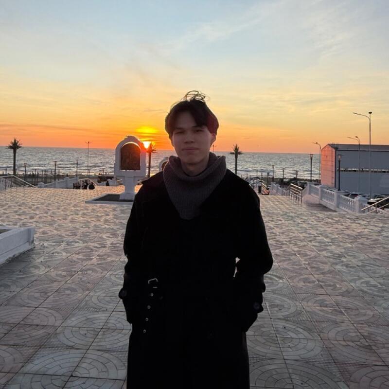 A young man in a black coat and gray scarf stands outside against a sunset backdrop near the sea.