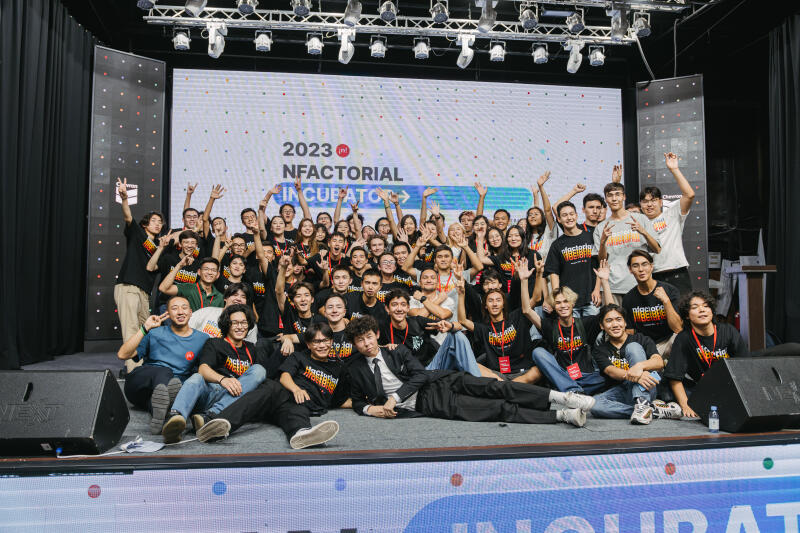 A large group of people is posing for a photo on a stage, with many raising their hands in the air. The backdrop features a screen displaying "2023 NFACTORIAL INCUBATOR".