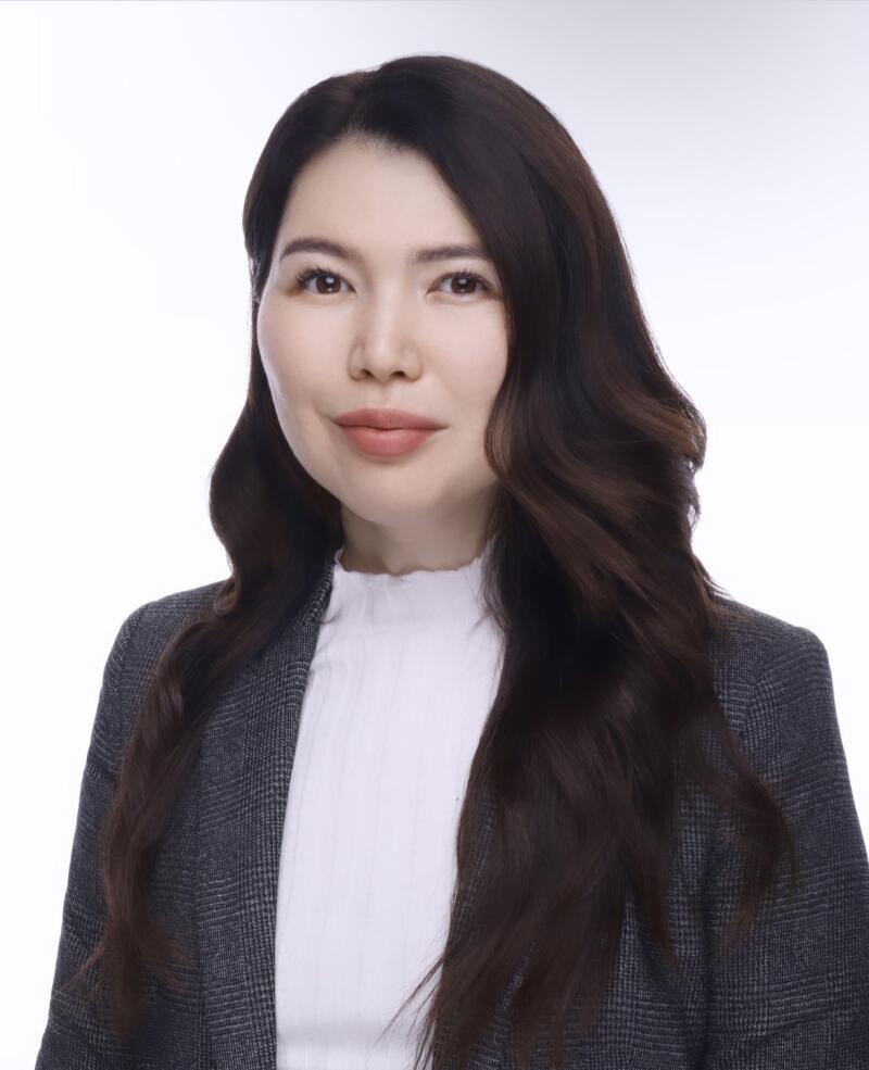 A woman with long, dark wavy hair and light skin wears a white top and a gray plaid blazer.