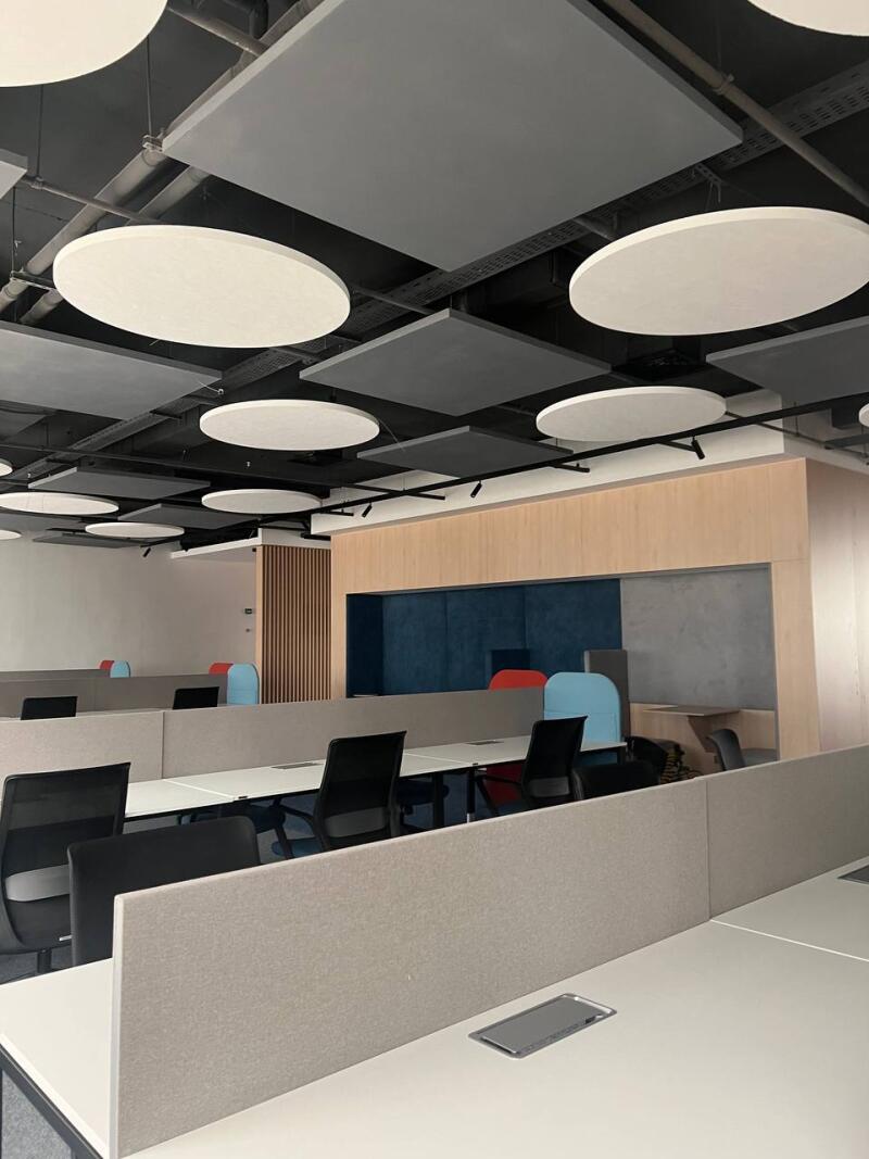 The photo shows an office space with rows of desks separated by gray partitions, black office chairs, and a ceiling with round and square acoustic panels.