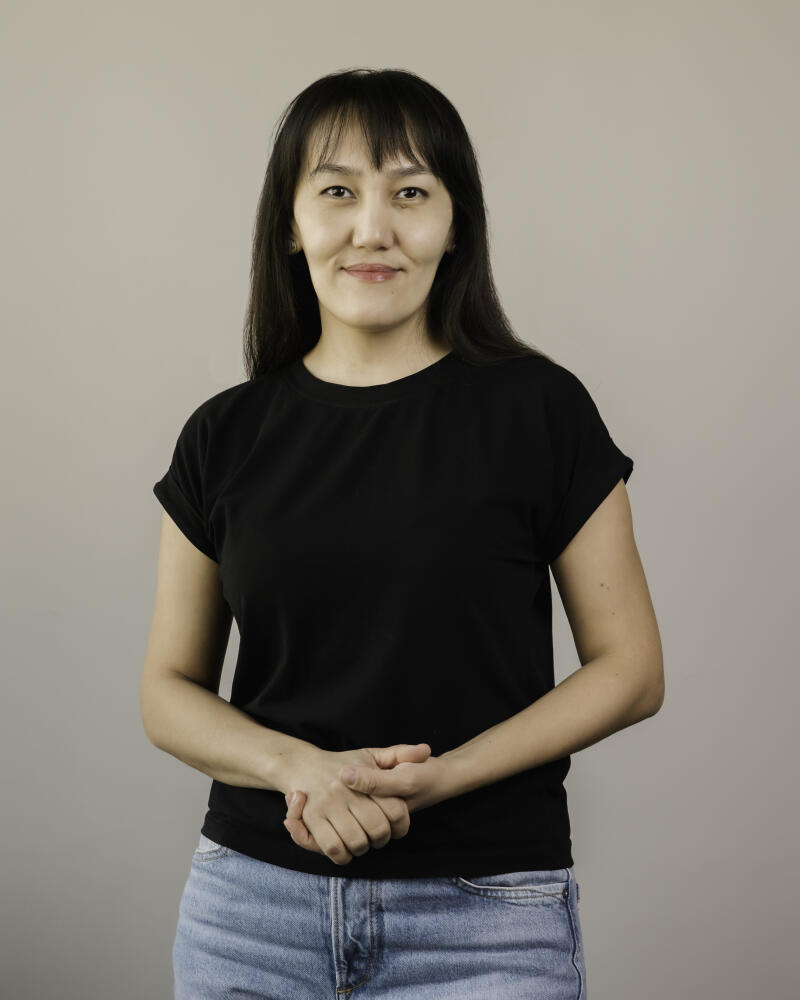 A woman with dark hair and bangs, wearing a black t-shirt and blue jeans, stands with her hands clasped.