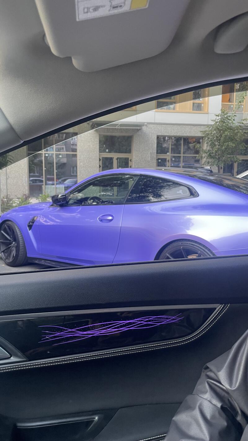 The image shows a lavender-colored car parked outside a building, as seen from inside another car with a door panel featuring decorative purple lines.