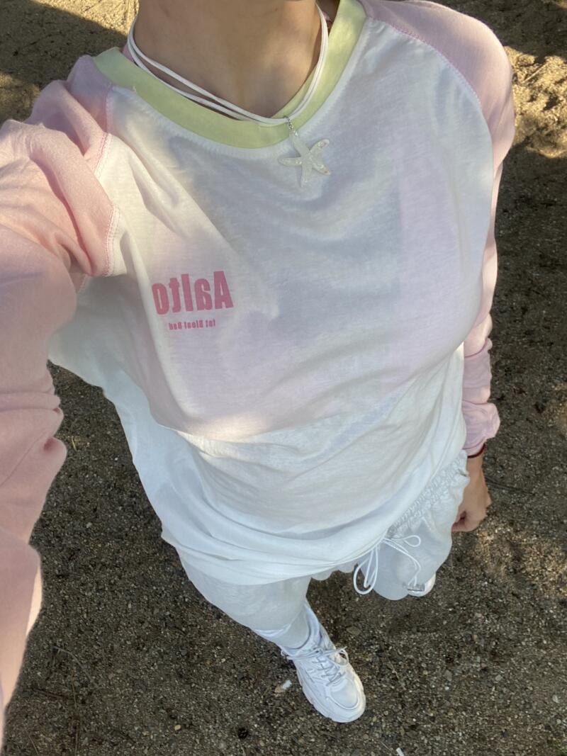 A person is wearing a white t-shirt with pink sleeves and the backwards text "Agisto" in pink, white shorts and white sneakers; they are also wearing a white necklace with a starfish charm.