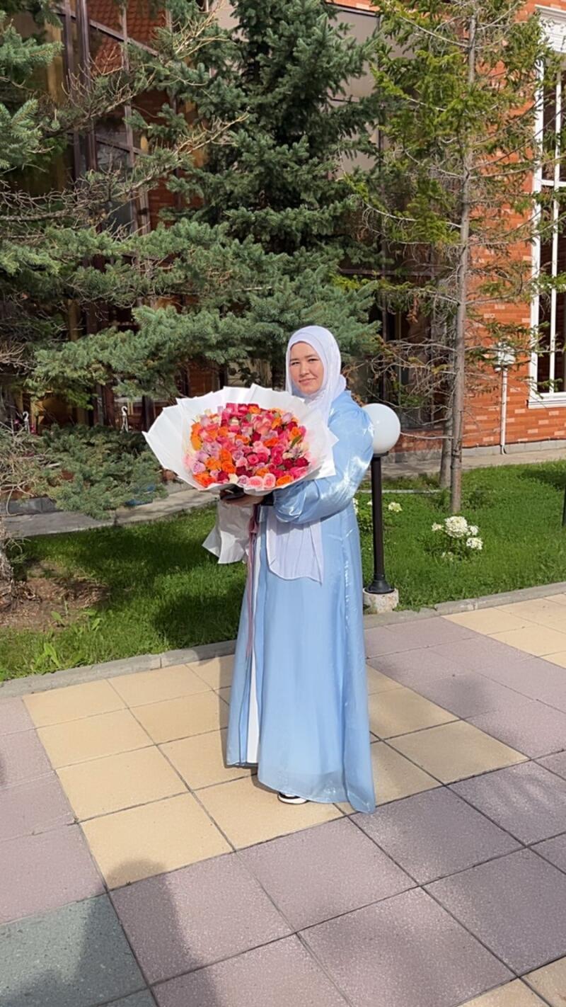 A woman wearing a hijab and a blue dress holds a large bouquet of roses in front of a building and trees.