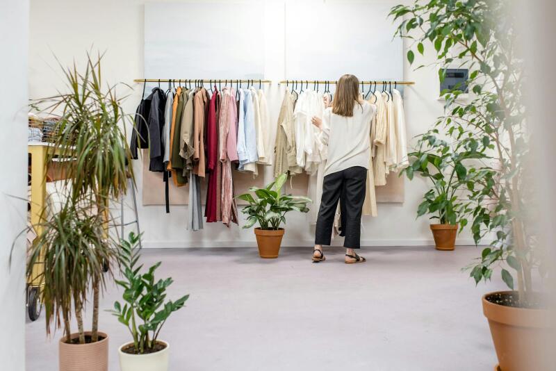 A person is looking at clothes hanging on a rack in a shop with several potted plants in the foreground and background.