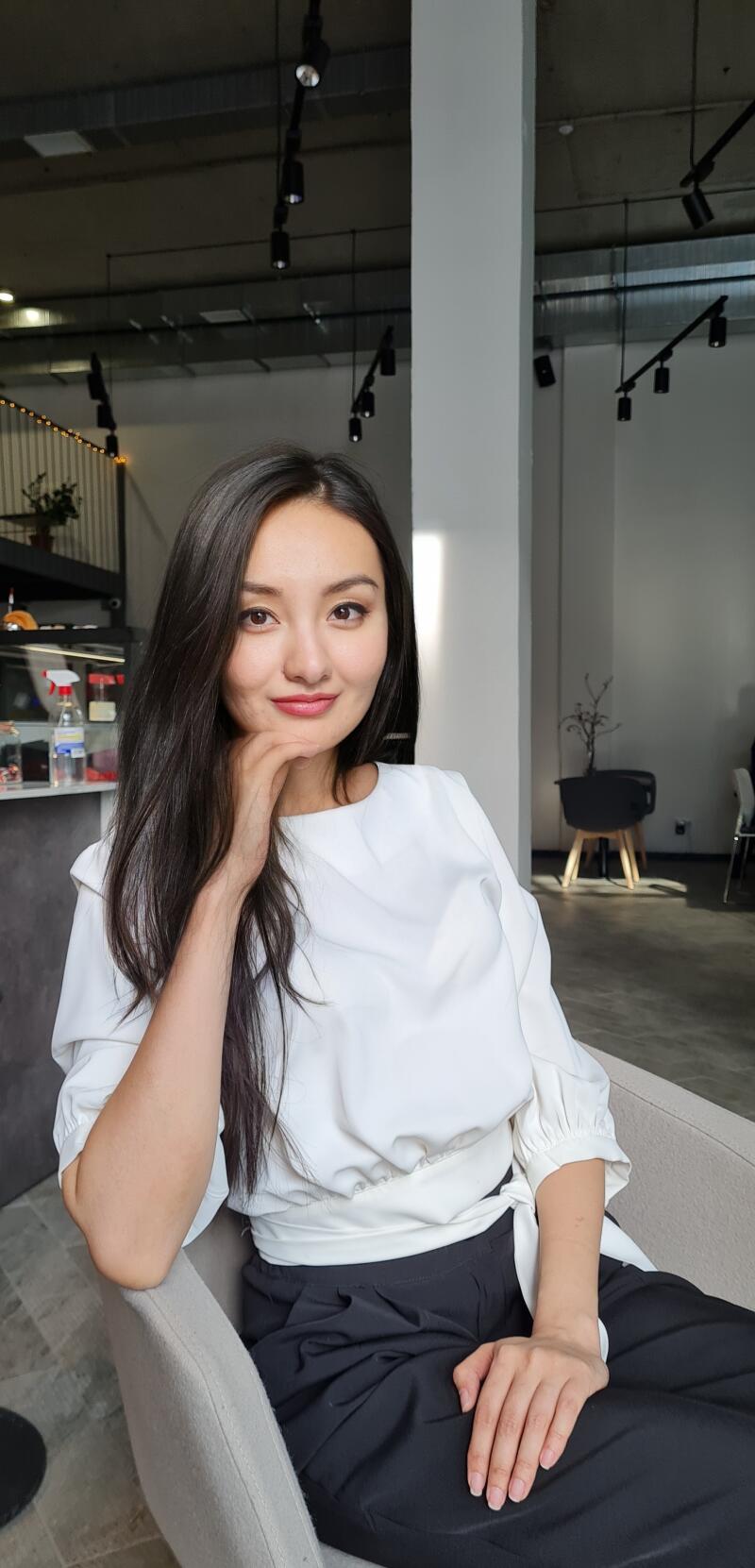 A woman with long, dark hair is sitting in a chair, wearing a white blouse and dark pants, with her hand on her chin.