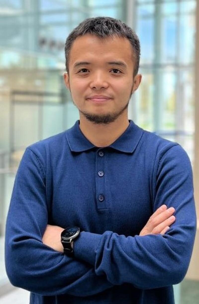 A man with short dark hair and a beard is wearing a blue long-sleeve polo shirt and a watch, standing with his arms crossed.