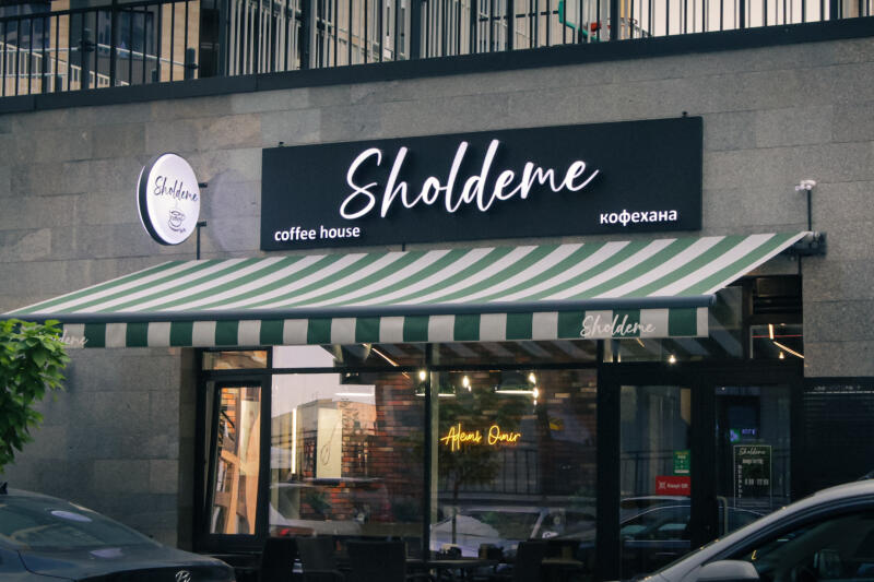 The exterior of a coffee house called "Sholdeme" features a striped green and white awning, black signage with the cafe's name in white script and the words "coffee house" and "кофехана", and a round sign with the cafe's name.