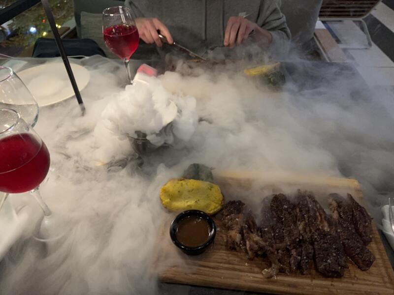 A person is cutting a steak surrounded by dense white smoke on a wooden serving board with a small bowl of sauce and yellow vegetable, alongside glasses of red liquid.