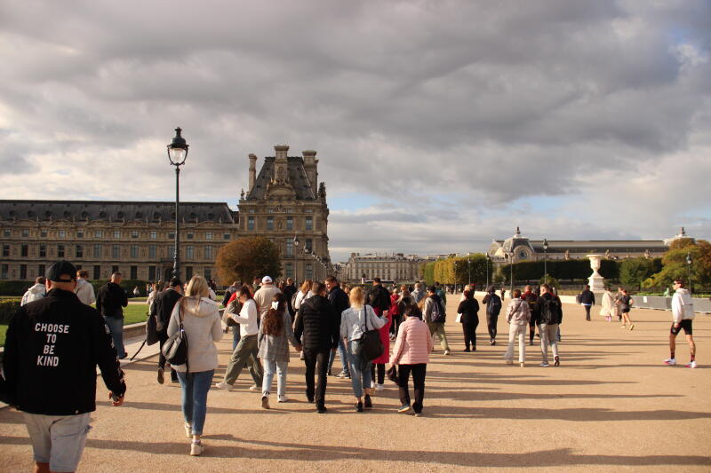 A crowd of people walk across a courtyard towards historic buildings under a cloudy sky; one man's jacket reads "CHOOSE TO BE KIND".