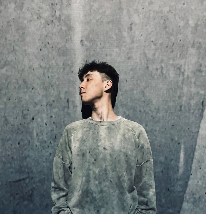 A person with dark hair and a gray sweatshirt stands in front of a gray wall, looking to the side.