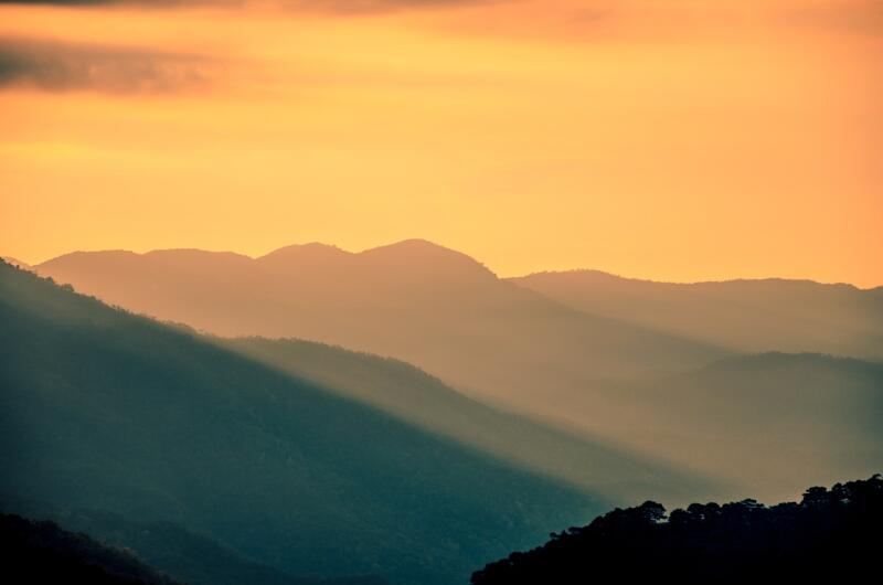 The image shows a landscape of layered mountains under a warm, orange-toned sky. Rays of light stream down the mountainsides, creating dramatic shadows.