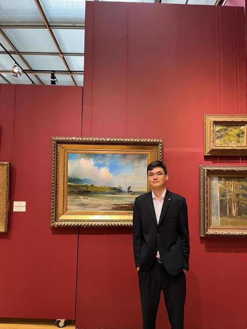 A man in a suit stands in front of art pieces on a red wall.