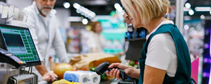 A cashier is scanning groceries at a checkout counter, where a customer is standing nearby.