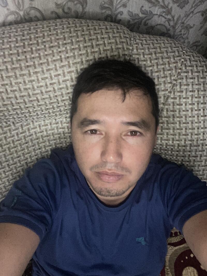 A selfie of a man with short dark hair and a mustache, wearing a blue t-shirt, lying on a patterned cushion.