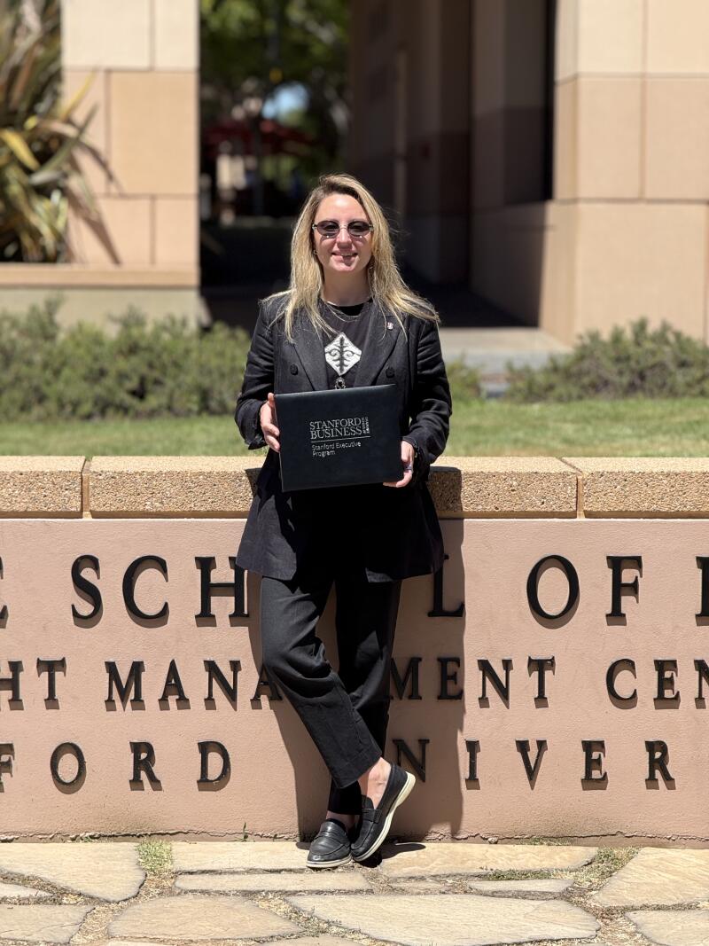 A woman in a black suit stands in front of a wall that reads "Stanford University" and holds a "Stanford Business" certificate.