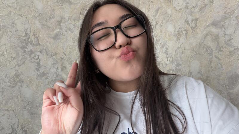 A young woman with long dark hair and glasses makes a kissing face with closed eyes, holding up two fingers in a peace sign. She wears a white t-shirt against a textured light-colored wall.