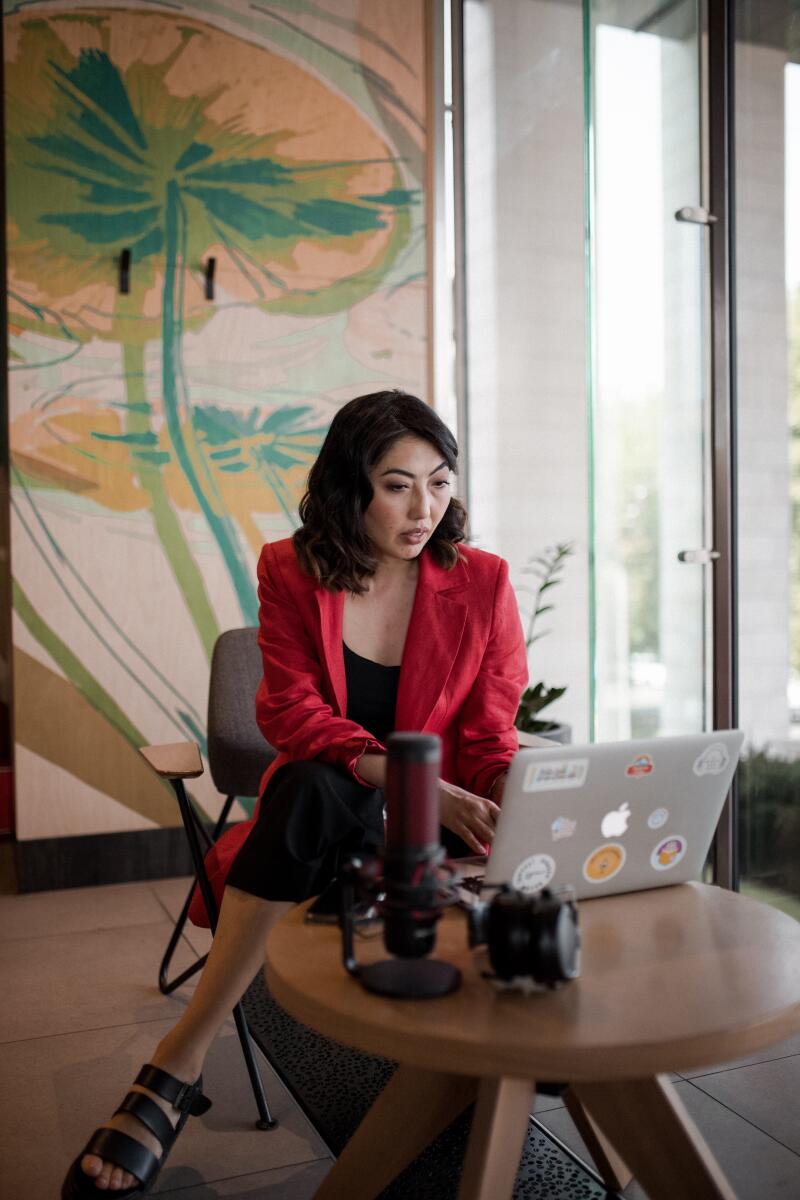 A woman with dark hair is sitting and working on a laptop near a microphone and headphones, wearing a red blazer and black sandals.