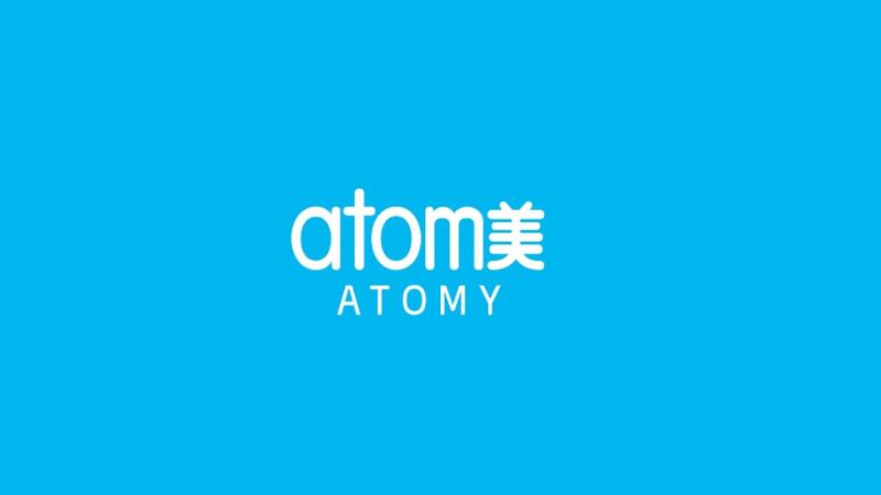 На изображении логотип "Atomy" белого цвета на синем фоне. Логотип состоит из слова "atom" строчными буквами, за которым следуют два китайских иероглифа, а под ними слово "ATOMY" заглавными буквами.