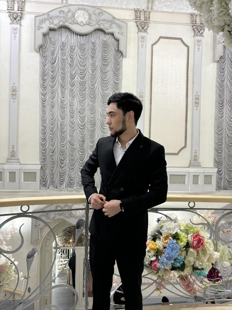 A man in a black suit stands on a balcony with a decorative railing, in front of a backdrop of ornate white walls and floral arrangements.