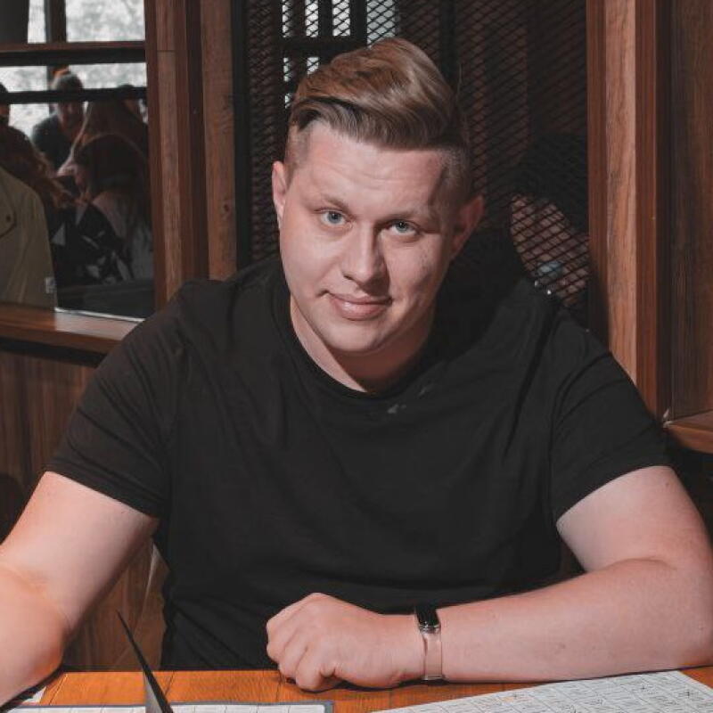 A man in a black t-shirt sits at a table, with a watch on his left wrist, and papers in front of him.