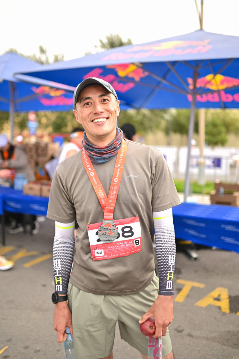 A smiling man in sportswear and a baseball cap is wearing a marathon finisher medal and a race bib that reads "Turkistan Marathon 2024". He is holding a water bottle and an apple.