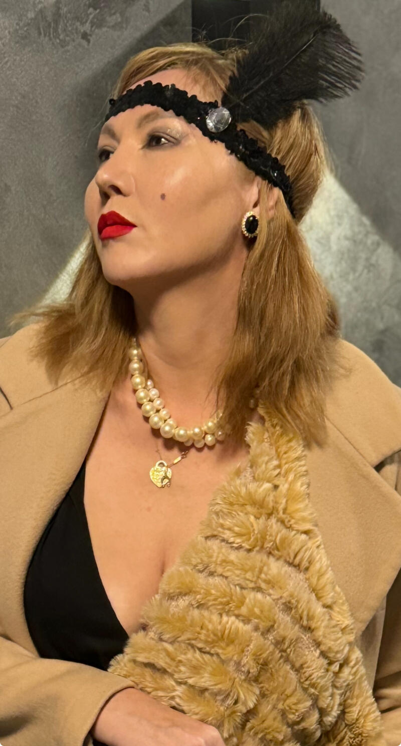 A woman in 1920s style, wearing a black headband with a feather and jewel, pearl necklaces, red lipstick, and a tan coat with fur trim.