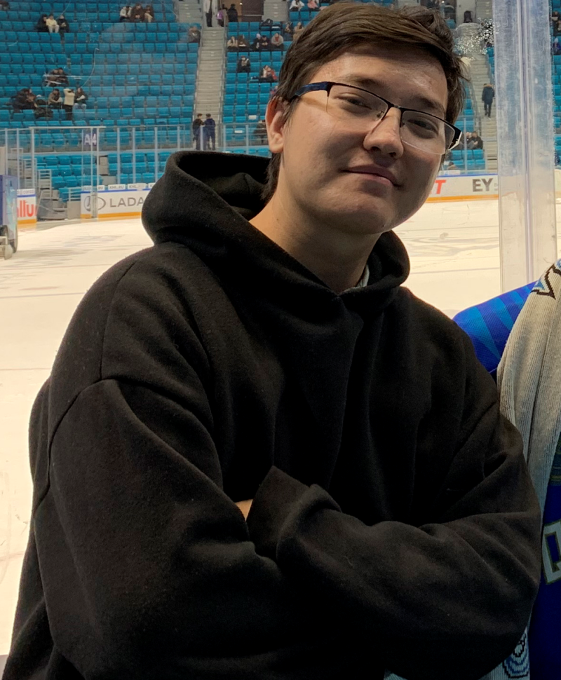 A person with glasses and a black hoodie stands in front of a hockey rink with blue stadium seating in the background.