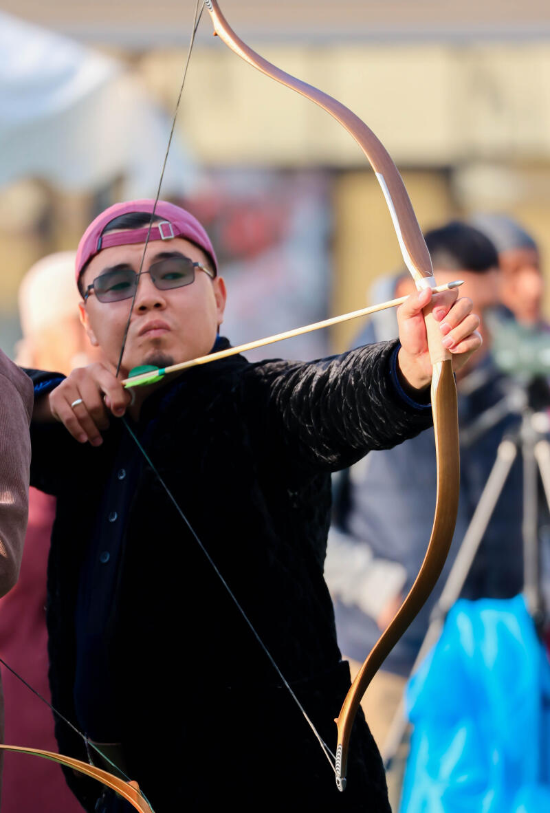 An archer in a pink cap and sunglasses aims with a recurve bow and arrow, holding the bow taut.