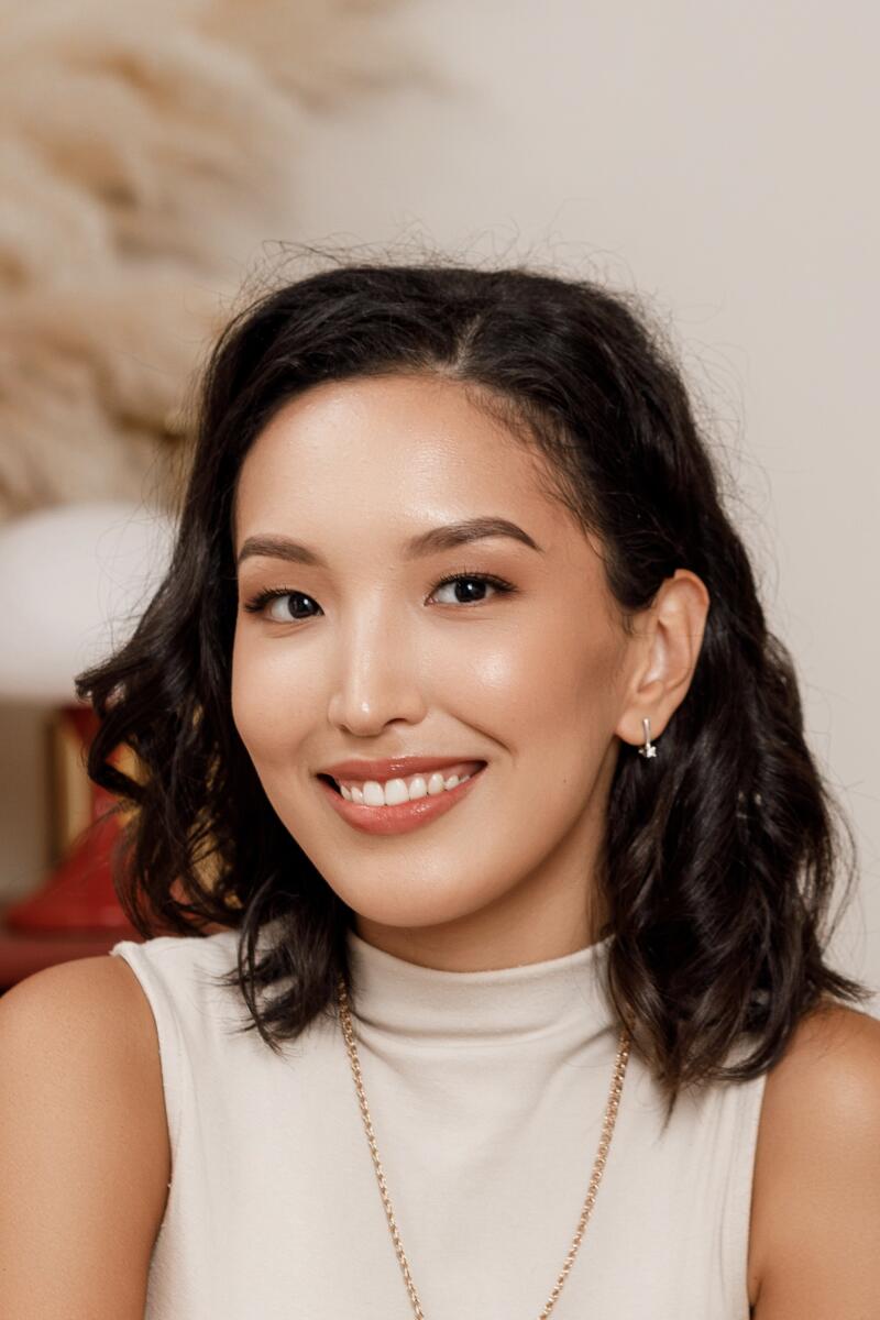 A young Asian woman with dark, wavy hair smiles at the camera, wearing a light-colored high-neck top, a gold chain necklace, and small star-shaped earrings.