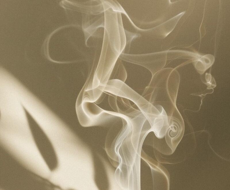 Light-colored smoke plumes curl upwards against a warm beige background, with shadows on the left and a smoke formation suggesting a face in profile on the right.