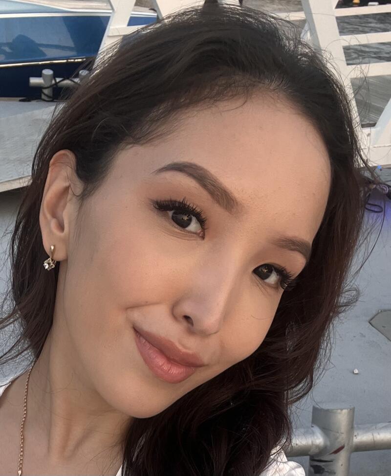 A close-up shot of a woman with dark hair, wearing a gold earring and necklace, with a boat visible in the background.