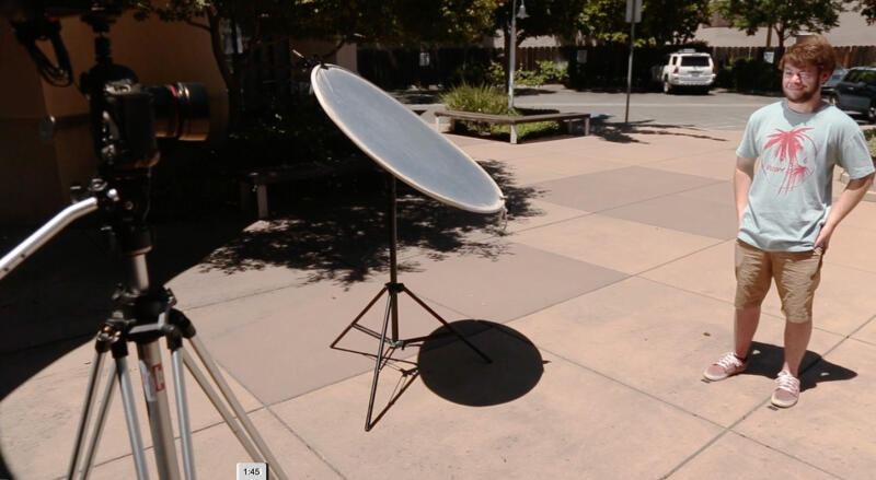 A man stands next to a photography reflector, with a camera on a tripod in the foreground.