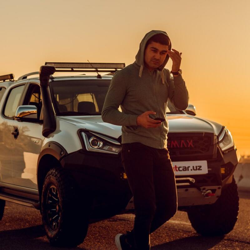 A man in a hoodie is standing next to a white off-road vehicle, looking at his phone and touching his forehead; the website 'tcar.uz' is visible on the vehicle's front.