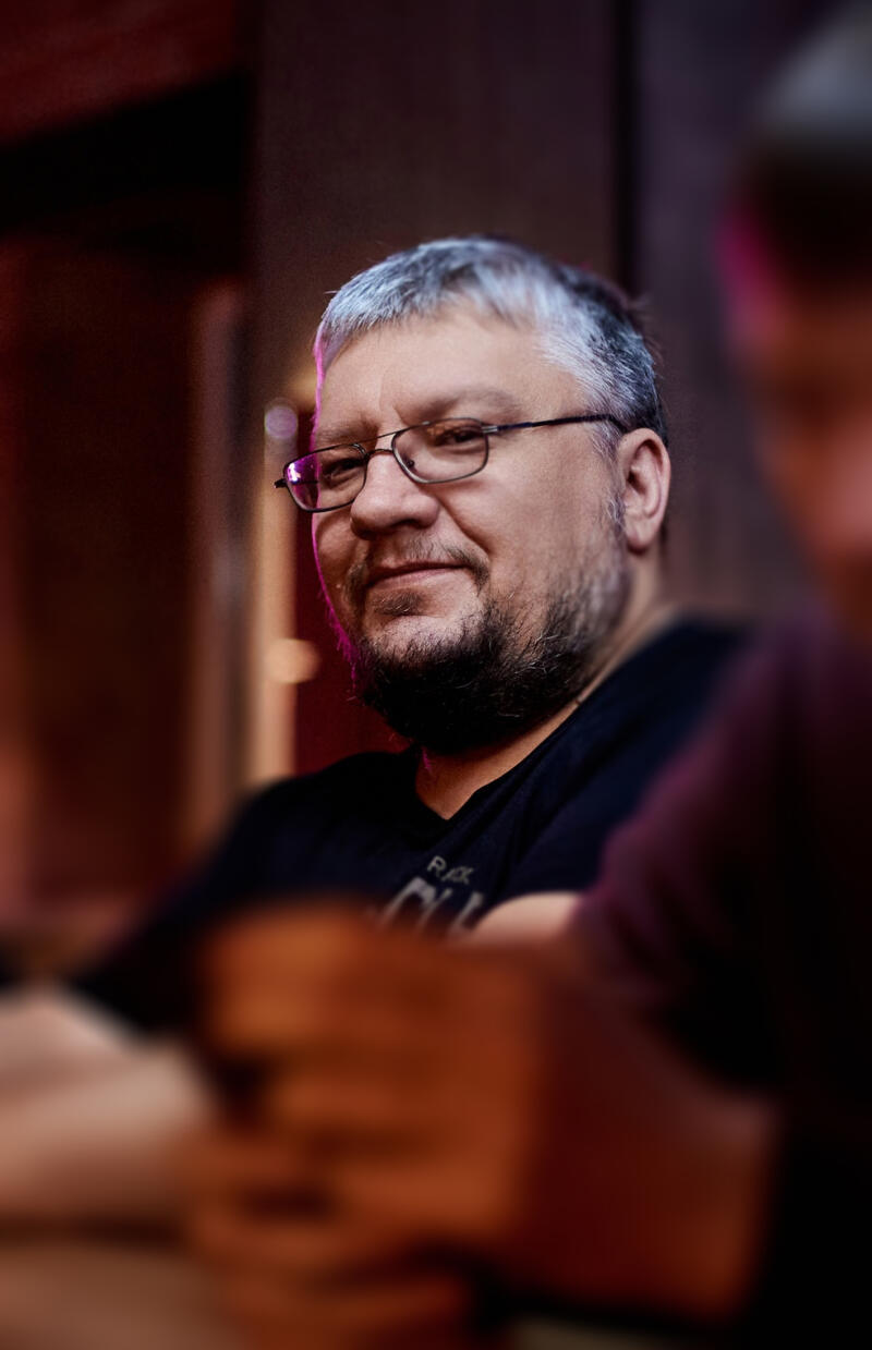 A middle-aged man with glasses and a beard is smiling slightly at the camera, wearing a dark shirt. The lighting is dim with a reddish hue.