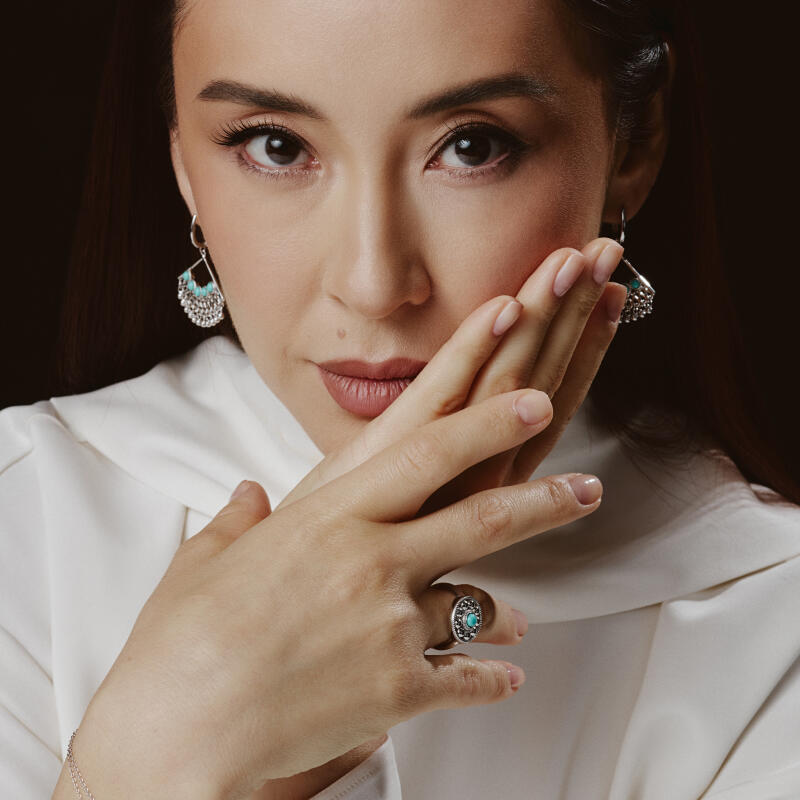 A woman with dark hair and fair skin is wearing silver earrings and a ring with a turquoise stone, posing with her hands near her face while wearing a white top.
