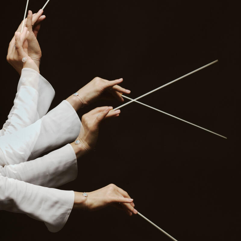 The image shows four arms in white long-sleeved shirts, each hand holding a conductor's baton against a dark background, with jewelry on the wrists and fingers.