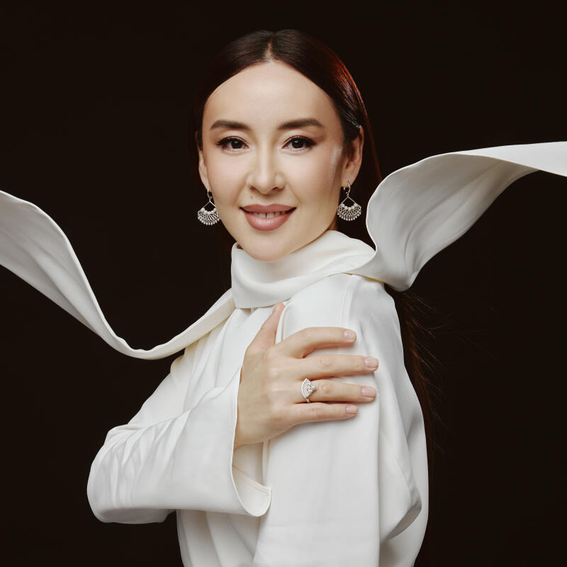 A woman with long dark hair is wearing a white dress and silver earrings and ring, posing against a dark background with a flowing white scarf.