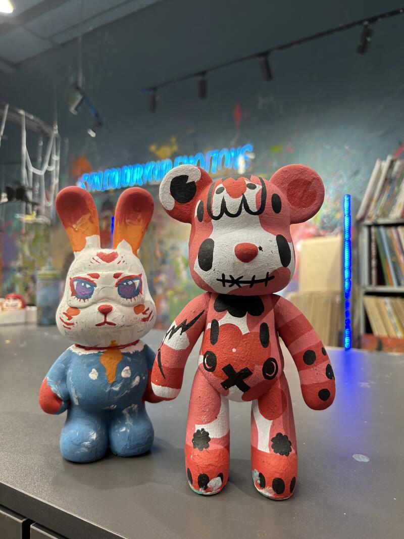A red and white bear-shaped figurine stands next to a blue and white rabbit figurine on a gray surface, with the words "RUN COLOUR YOUR EMOTIONS" visible in the background.