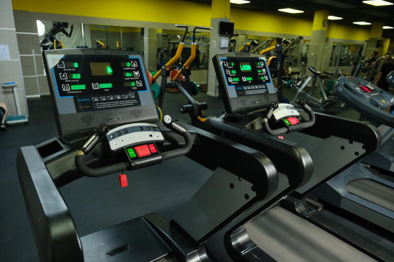 The image shows two treadmills in a gym setting, featuring digital displays and control panels with buttons. Various other exercise machines and mirrors are visible in the background.