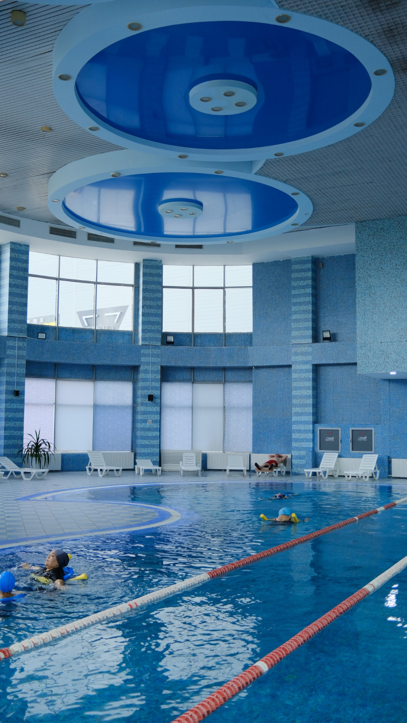 The image shows an indoor swimming pool with multiple swimmers, lane dividers, and lounge chairs along the side, under a blue-themed ceiling with circular designs.