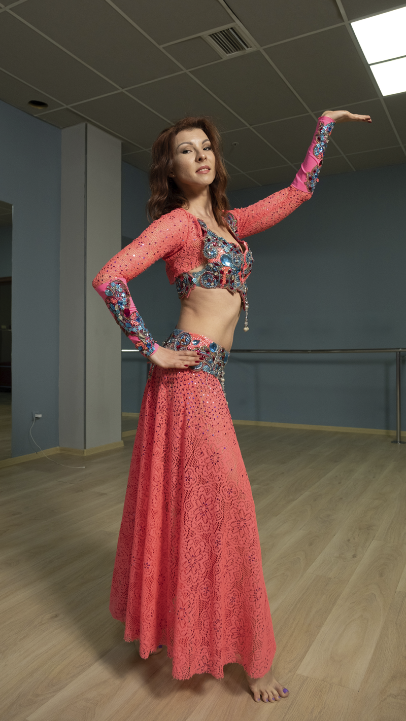 A woman in a coral-colored belly dancing outfit with blue and silver accents poses with one arm raised.