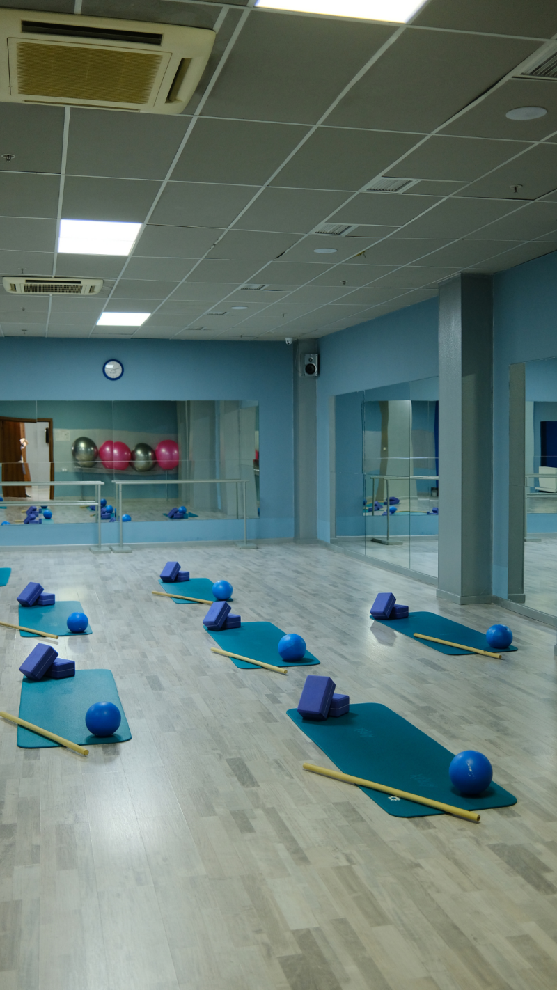 A fitness studio with blue walls and light wood-patterned floors features mats, yoga blocks, exercise balls, and wooden rods arranged in rows.