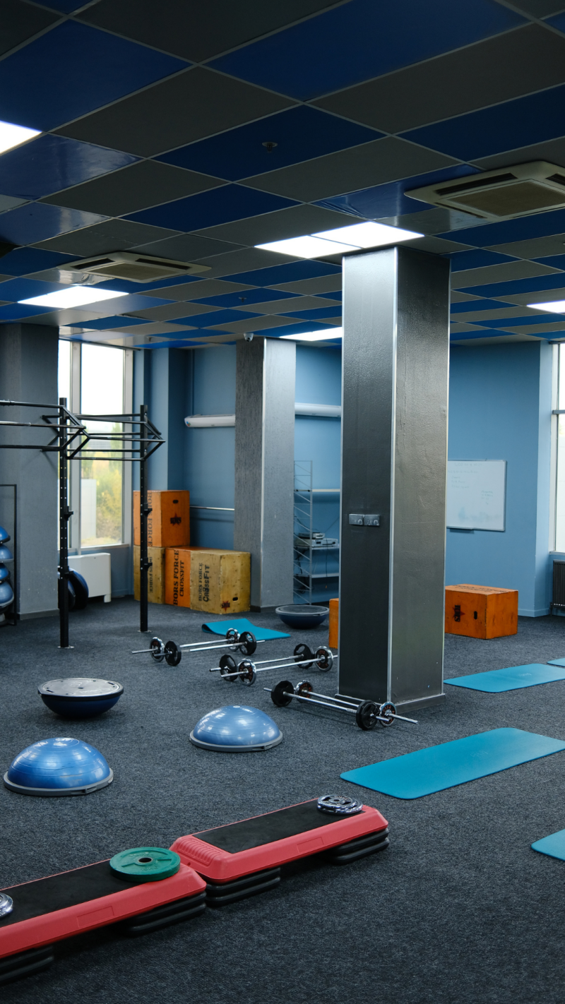 The interior of a gym features equipment such as barbells, BOSU balls, and step platforms, set against blue walls and a checkered ceiling.